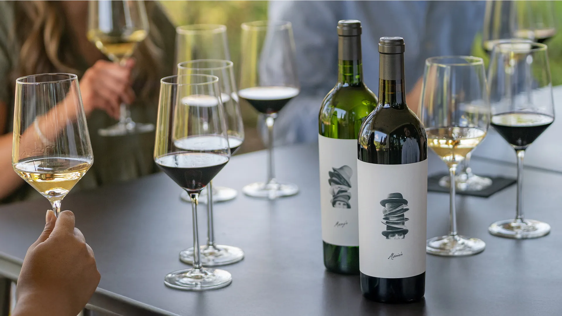 Several glasses of white and red wine with two wine bottles on a table, surrounded by people holding wine glasses at events.