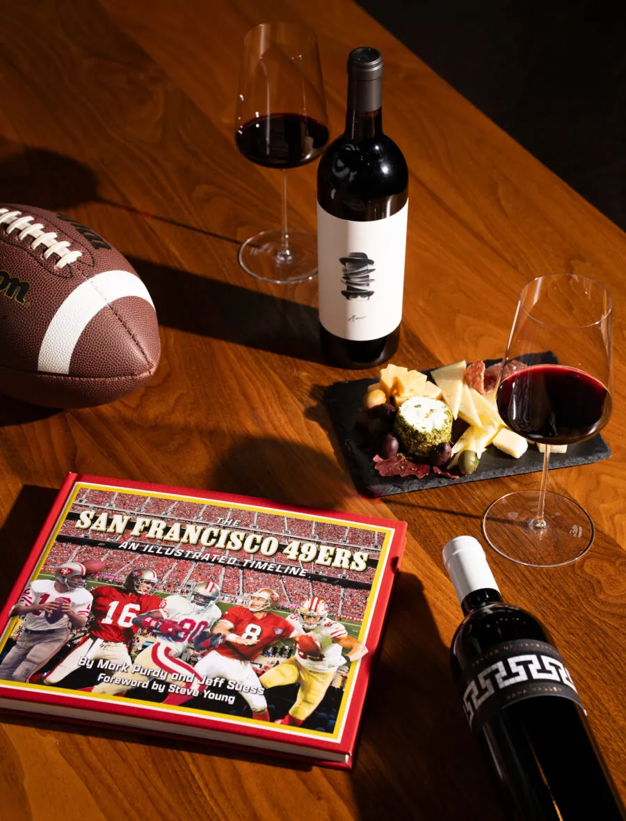 A football, two glasses and bottles of red wine, a cheese and charcuterie board, and a San Francisco 49ers book set the perfect scene for memorable events on a wooden table.