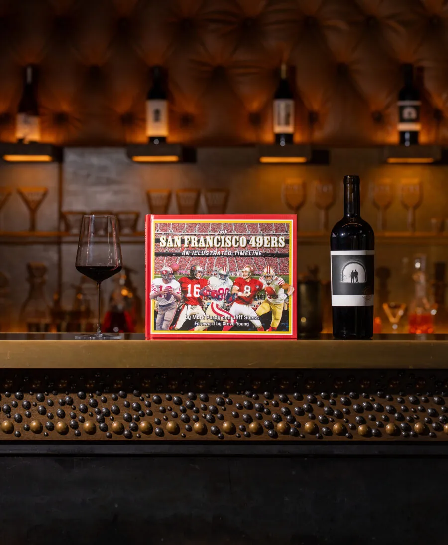 A box featuring San Francisco 49ers football players is displayed on a bar counter beside a glass and a bottle of red wine, creating the perfect backdrop for sports-themed events, with shelves and bottles in the background.