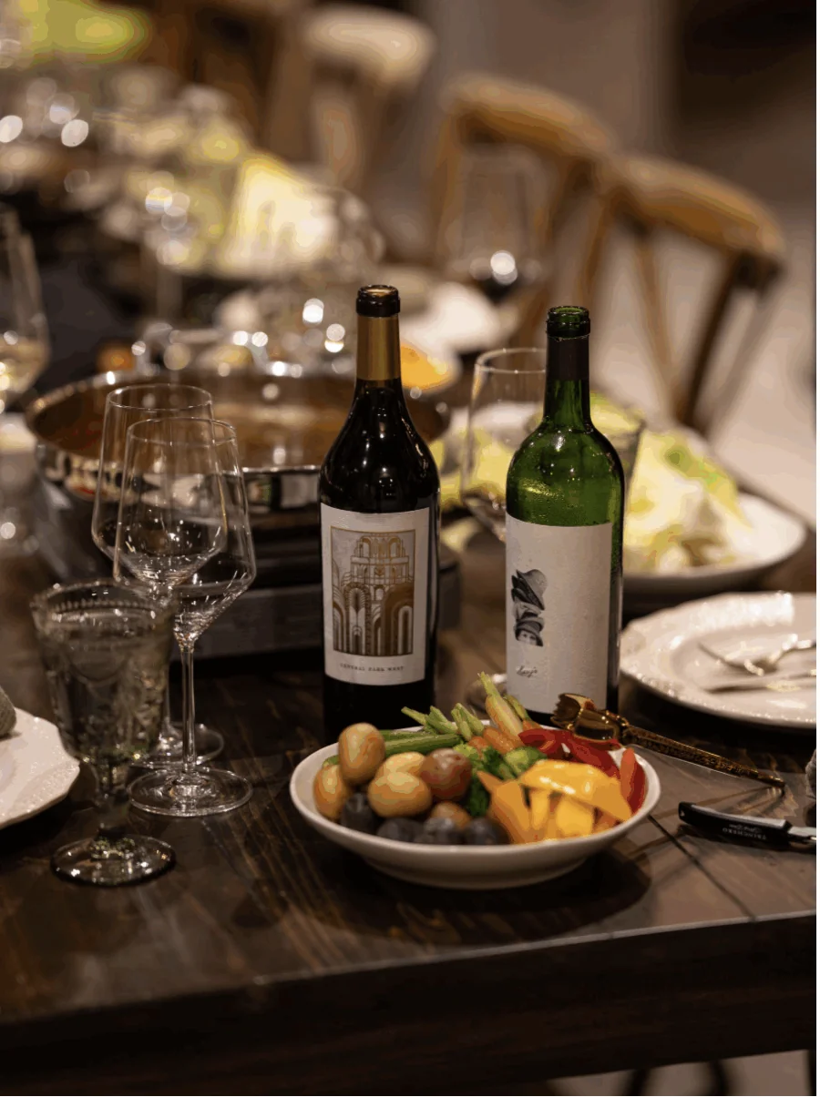 A dining table set for events, featuring wine bottles, empty glasses, a bowl of mixed vegetables and potatoes, plates, and cutlery, all prepared for a meal.