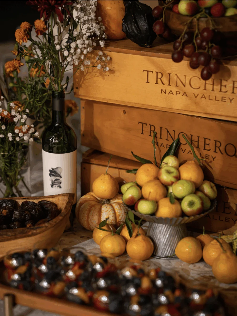 A display for events features fresh fruits, dates, a bottle of wine, flowers, and wooden crates labeled 