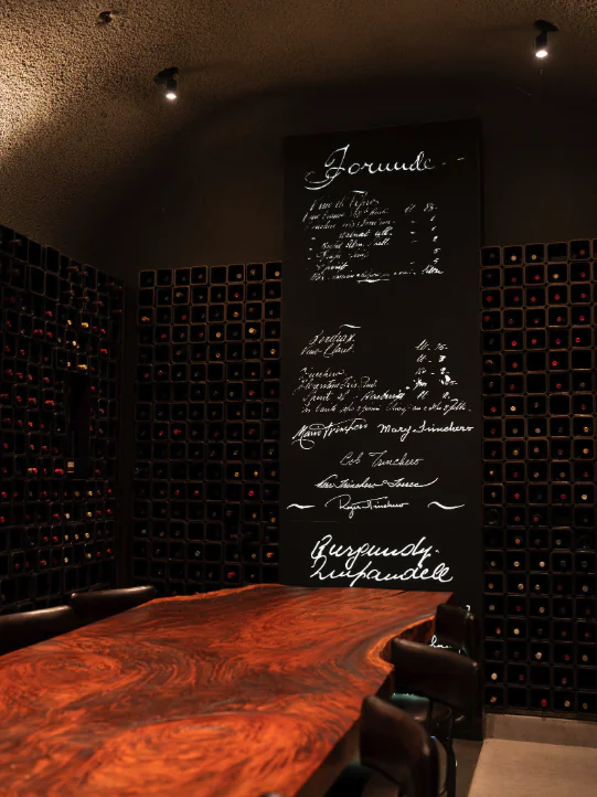 A wooden table and chairs are set in a wine cellar with walls of wine bottles, a large chalkboard menu in cursive script, creating an inviting setting for Napa Valley Events.