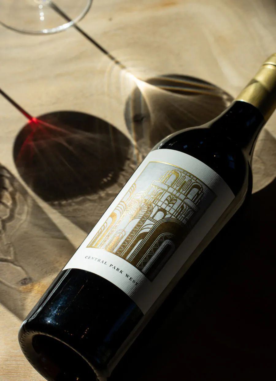 A bottle of Central Park West red wine lies on a wooden table beside a wine glass casting a red-tinted shadow—perfect for savoring after attending Napa Valley Events.