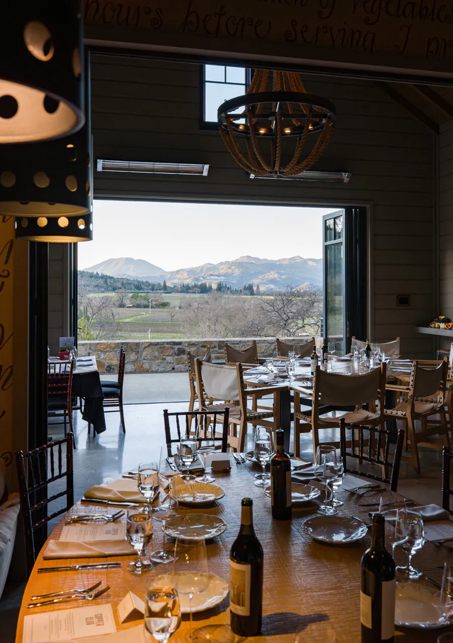 A dining room with set tables and wine bottles, looking out through open doors to a patio and a scenic view of mountains and vineyards—perfect for unforgettable Napa Valley Events.
