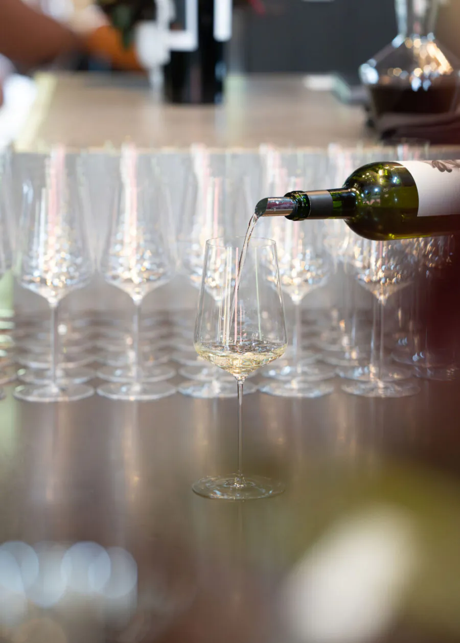 A bottle pours white wine into a glass, with several empty wine glasses lined up in the background on a reflective countertop—setting the stage for unforgettable Napa Valley Events.