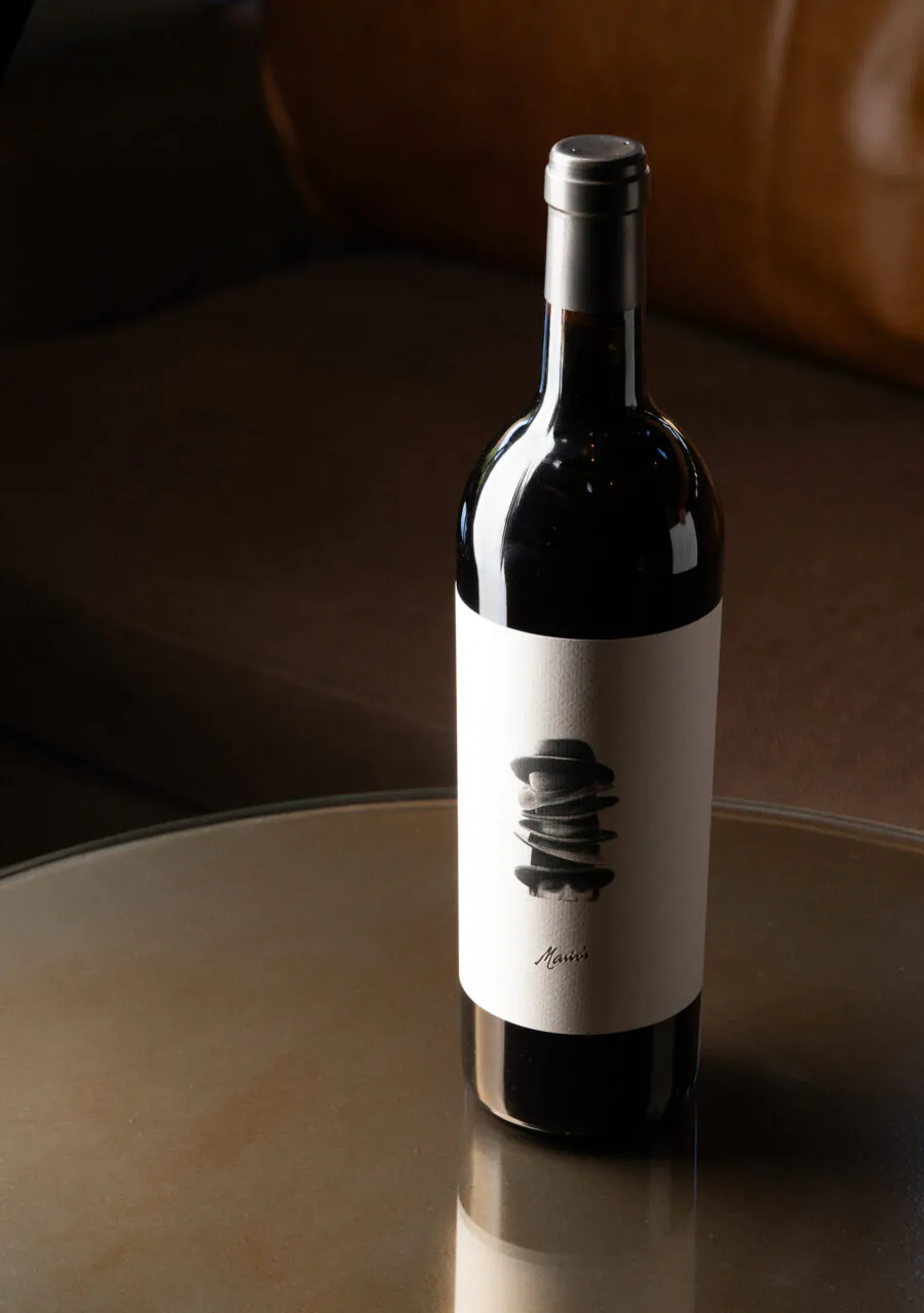 A bottle of red wine with a minimalist label sits on a round glass table, evoking the relaxed elegance of Napa Valley Events, with a brown couch in the background.