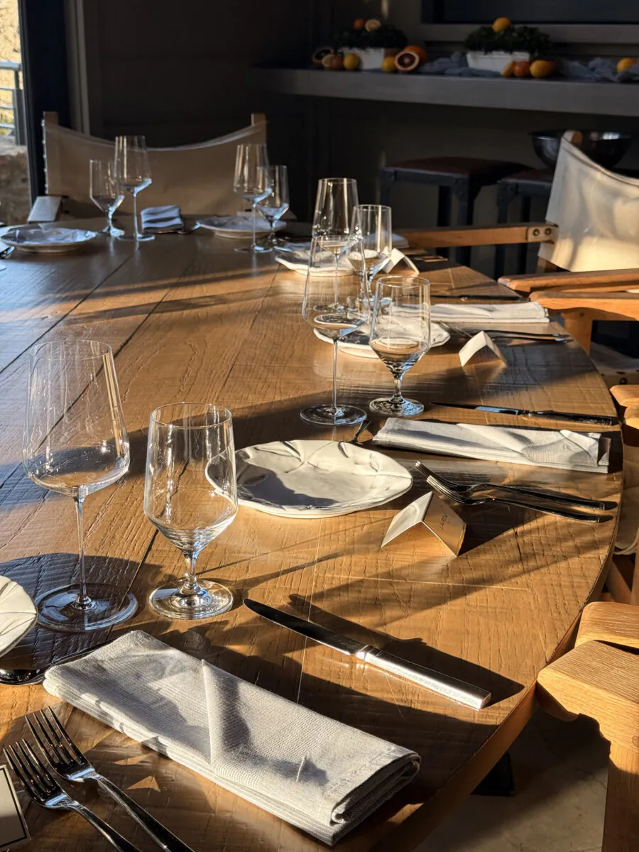A sunlit wooden table is set for six with plates, glasses, cutlery, and napkins—perfect for intimate Napa Valley events. Empty chairs surround the table; fruit and glasses are visible in the background.